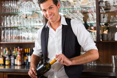 Barman standing behind the bar In restaurant opening a wine bottleの写真素材