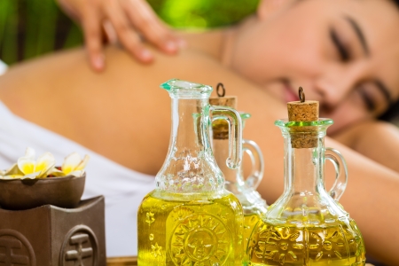 Beautiful Asian woman having a wellness back massage in a tropical setting and feeling visibly good about it - Essential oils are in the foregroundの写真素材