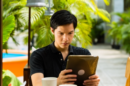 Asian man is sitting in a bar or cafe outdoor and is surfing the internet with a tablet computerの写真素材