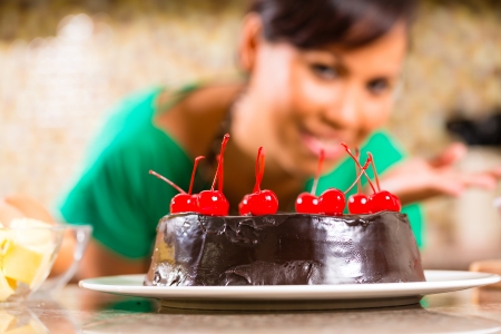 Asian woman presenting homemade chocolate cake with cherries she baked in her kitchen for dessertの写真素材