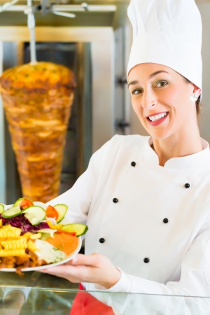 Doner kebab - friendly female vendor in a Turkish fast food eatery, holding plate with fries and kebab in front of skewerの写真素材