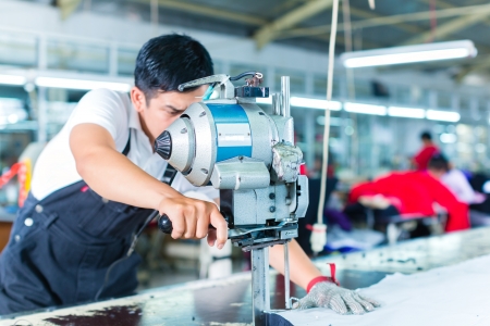 Indonesian worker using a cutter - a large machine for cutting fabrics - in a asian textile factory, he wears a chain glove Indonesian worker using a cutter - a large machine for cutting fabrics - in a asian textile factory, he wears a chain glove