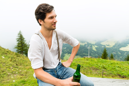 Hiking - Young man sitting at mountain hut with beer bottle in the Bavarian Alps enjoying the panorama in the leisure time or in vacationの写真素材