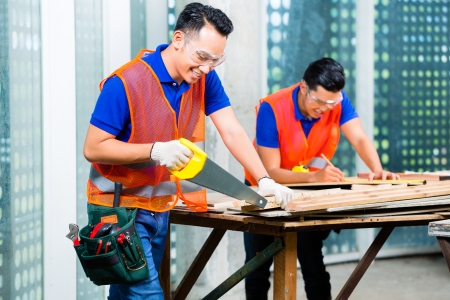 Asian Indonesian builder or craftsman sawing with a saw a wood board of a tower building or construction site wearing protection glasses and glovesの写真素材