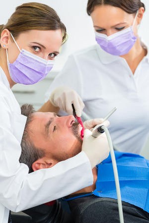 dentist in her practice or office treating male patient with assistant wearing masks and glovesの写真素材