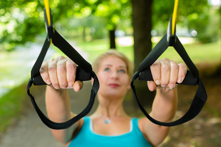 Young woman exercising with suspension trainer sling in City Park under summer trees for sport fitnessの写真素材