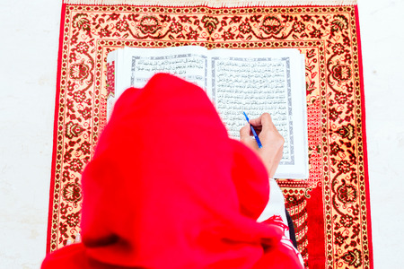 Asian Muslim woman reading Koran or Quran on praying carpet wearing traditional dressの写真素材