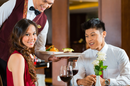 Asian Chinese couple - Man and woman - or lovers having a date or romantic dinner in a fancy restaurant while the waiter is serving foodの写真素材
