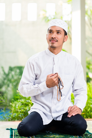 Asian Muslim man praying at home sitting on prayer carpet in his house in front of the tropical gardenの写真素材