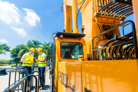 Asian construction machinery driver discussing with foreman blueprints on pad or tablet computer of...