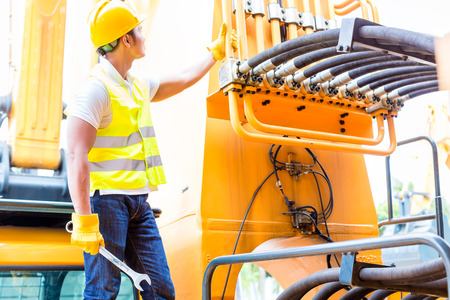 Asian motor mechanic standing in front of construction or mining machinery in vehicle workshop