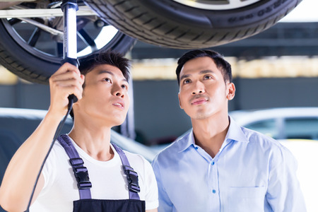 Asian Chinese car mechanic with customer looking at auto floor with lampの写真素材