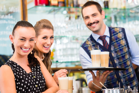 Two woman friends in coffee bar drinking latte macchiato in glass, a barista preparing drinks in the backgroundの写真素材
