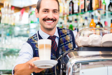 Barista in coffee bar offers latte macchiato in glass, in the background bottlesの写真素材