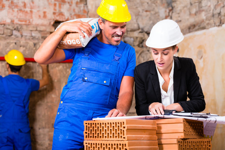 Architect and construction worker having sack of cement on shoulder on site with planの写真素材
