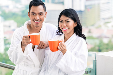 Asian couple in morning front of city skyline drinking coffee still in bathrobeの写真素材