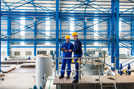 Two workers in large metal workshop or factory checking work standing on large machine Two workers in large metal workshop or factory checking work standing on large machine