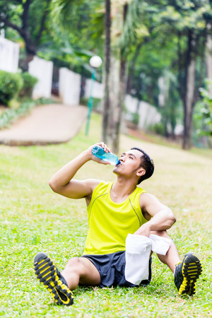 Asian man having break from sport training in tropical park, drinking water from a bottleの写真素材