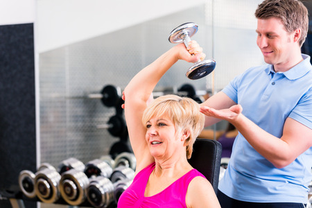 Senior woman at sport exercise with dumbbell in gym with trainer to gain strength and fitnessの写真素材