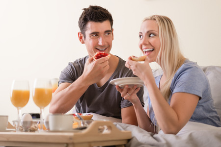 Happy playful couple enjoying breakfast in bed grinning at each other as they prepare to bite into toast and jam with a spread of orange juice, coffee and eggs on a tray in front of themの写真素材