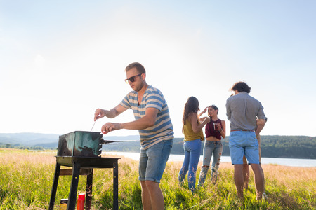 Gang of young men and women having barbecue at lake in summerの写真素材