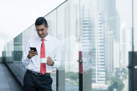 Indian businessman drinking coffee and using a smartphone on the terrace of a modern buildingの写真素材