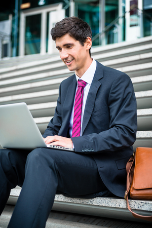 Young businessman using a laptop with wireless internet connection while sitting down on stairs outdoorsの写真素材
