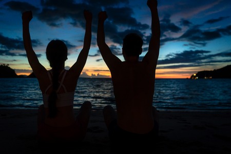 Rear view of a young couple sitting on the beach with raised arms while feeling happy and free during summer vacation in Flores Island, Indonesiaの写真素材