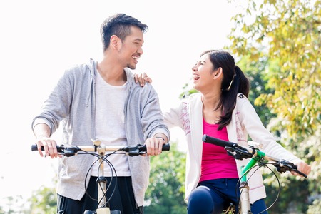 Young Asian couple laughing together while riding bicycles outdoors in summerの写真素材