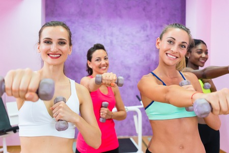 Determined beautiful women smiling while exercising with dumbbells for toned arms during group class in a contemporary fitness clubの写真素材