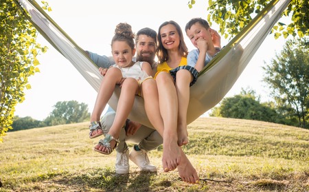Family portrait with a beautiful mother of two playful children swinging in a hammock while looking at camera next to her husband outdoors in summerの写真素材