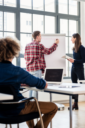 Rear view of a student writing on a training paper board while presenting with his classmate a new lesson, in front of the class at a modern college or universityの写真素材