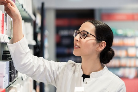 Side view close-up portrait of a dedicated female pharmacist standing in front of the pharmacy shelves, with various products while thinking to make the best choiceの写真素材