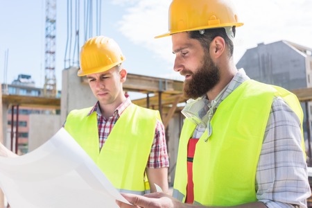 Two young construction workers wearing yellow hard hats and reflective safety vests while analyzing...