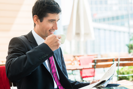 Young businessman smiling while reading a newspaper and drinking coffee in a cafeteria in the morningの写真素材