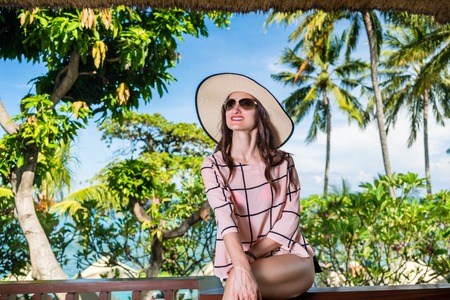 Woman wearing straw hat and sunglasses, sitting in front of palm trees in summer holidaysの写真素材