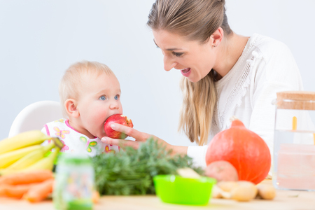 Portrait of a cute and healthy baby girl looking with curiosity at the pink fruit puree, while sitting on a high chair during meal with her happy mother at homeの写真素材