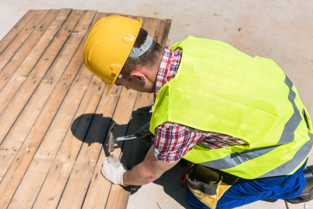 High-angle view of a blue-collar worker wearing yellow hard hat, safety vest and gloves while using a hammer during work on the construction siteの写真素材