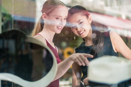 Two women friends shopping in a fashion boutique pointing to a display of trendy hat viewed through the glass with reflectionsの写真素材