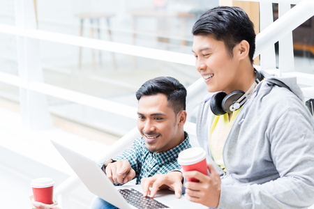 Two young Asian men using a laptop while relaxing during break on the stairs of a modern buildingの写真素材