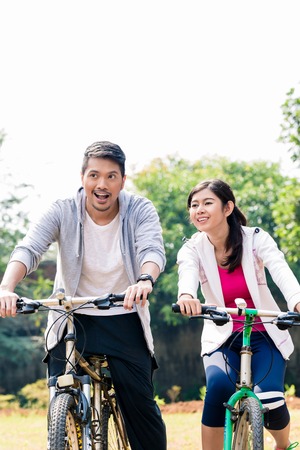 Young Asian couple laughing together while riding bicycles outdoors in summer