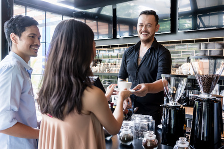 Cheerful bartender serving coffee over the bar counter to a young female customer standing next to her boyfriend in the cafeteriaの写真素材