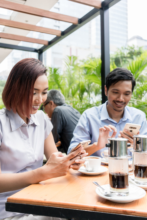 Three young Asian friends smiling while using electronic devices connected to the wireless internet network of a modern coffee shopの写真素材