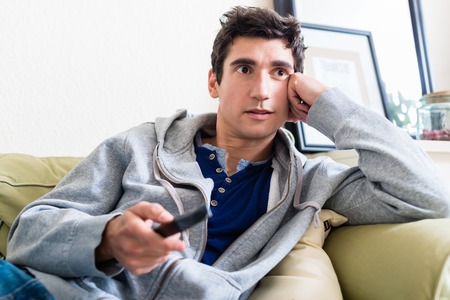 Portrait of young man sitting on the sofa with the remote control in his hand while watching TV at homeの写真素材