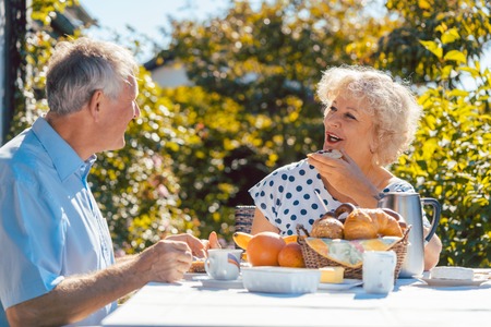 Senior woman and man having breakfast sitting in their garden outdoors in summer, eating bread rolls and drinking coffeeの写真素材
