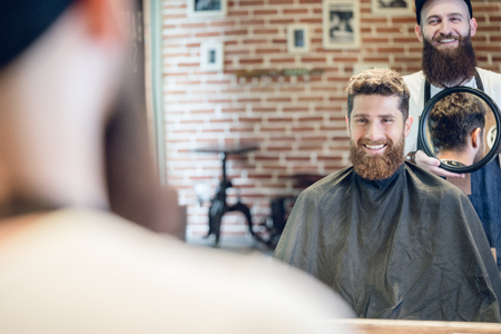 Over the shoulder view of a handsome young man smiling, while looking at his new trendy haircut in the mirror held by his experienced barber in a cool hair salonの写真素材