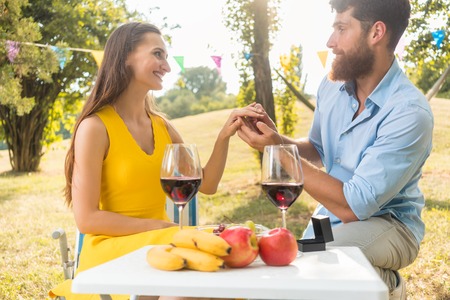 Young handsome man sitting on bended knee while showing to his beautiful girlfriend an engagement ring during romantic picnic with red wine and fresh fruitsの写真素材
