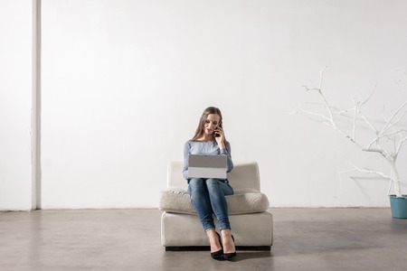Young woman sitting inwhite room using smartphone and notebook PCの写真素材