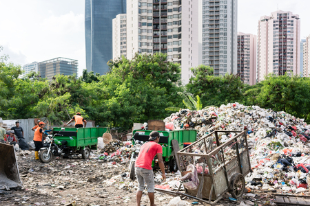 People disposing waste materials and garbage in a landfill site next to a modern district with skyscrapers in Jakartaの写真素材