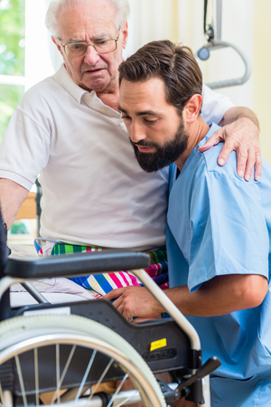 Elderly care nurse helping senior from bed to wheel chair in hospital or nursing home